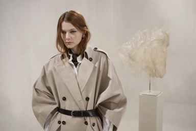 Model Louise Robert posing in an oversized beige double-breasted trench coat with a black belt next to a white sculpted art piece.