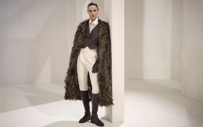 Model Cate Underwood leans against a white architectural column wearing a shaggy coat, tailored vest, white pants, and tall riding boots.