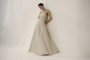 Model with short dark hair leaning against an off-white textured wall wearing a strapless sparkling ivory evening gown.