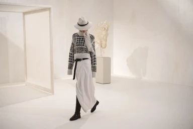 Model wearing a patterned knit sweater, wide-brimmed hat, long white skirt, and black boots walking across a white minimalist set.