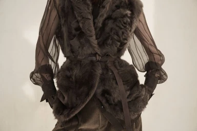 Model wearing a dark brown textured shearling vest with waist belt over sheer sleeves and corduroy trousers.
