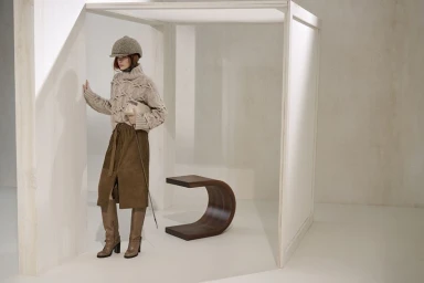 Model wearing a cable knit sweater, suede skirt, tall boots, and cap in a minimalist architectural studio set.
