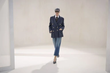 Model wearing a navy double-breasted blazer, cropped denim jeans, cap, sunglasses, and heels walking across a pale studio set.