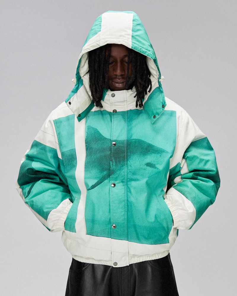 An adult in a hooded puffer jacket with a white and teal abstract print poses in front of a light gray background.