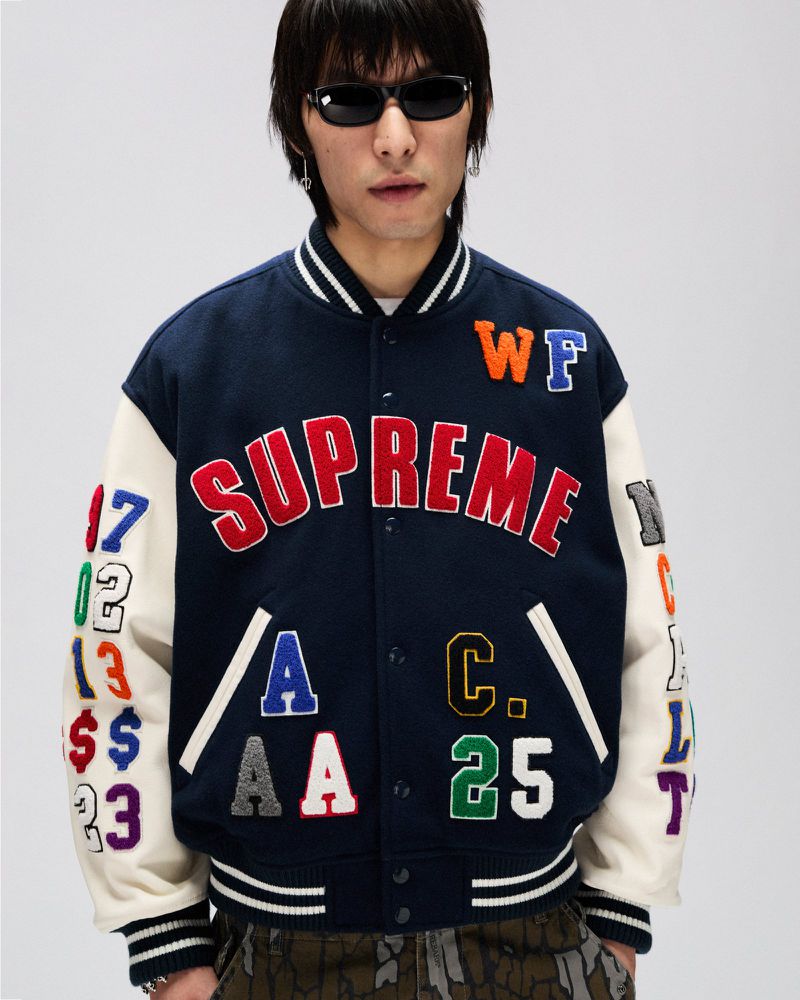 Studio shot of a man in a Supreme varsity jacket with colorful patch details, styled with dark sunglasses, against a bright backdrop.