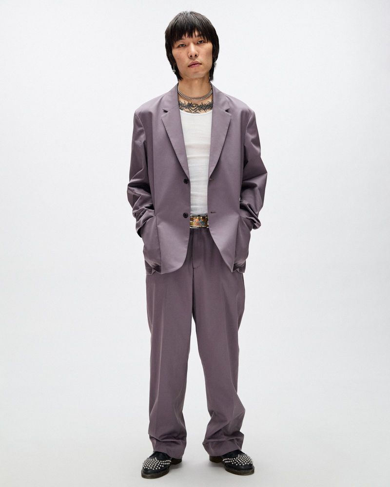 Full-length shot of a slender Asian male in an oversized mauve suit, white tank top, decorative belt, and black studded shoes on white.
