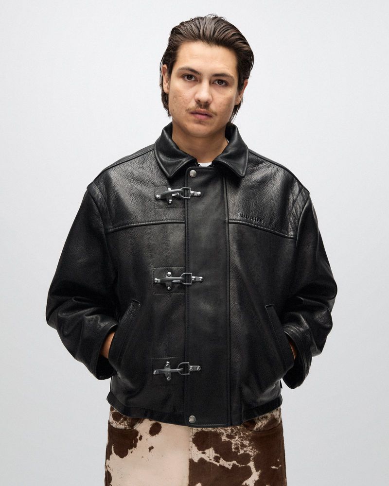Man poses wearing a black leather jacket with metal clasps, styled with a cow-print skirt, against a simple neutral background.