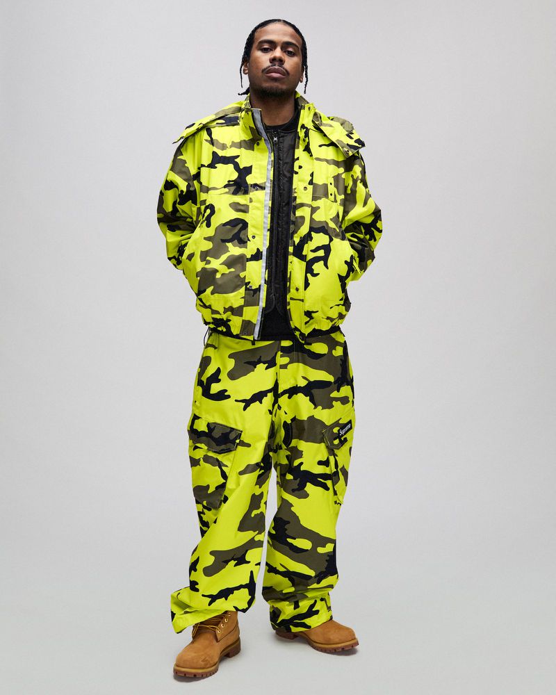 Adult male in a full-length studio shot wearing a yellow and black camouflage jacket and pants, paired with brown work boots.
