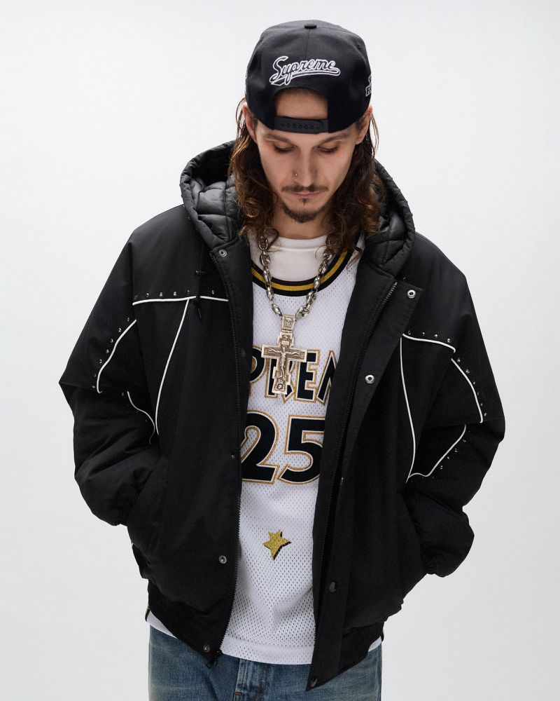 Man in black hooded jacket and baseball cap with silver chain posing indoors against a white wall, wearing basketball jersey.