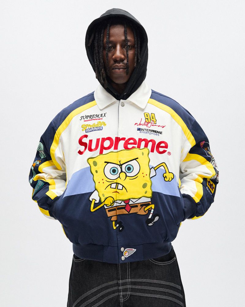 Adult male wearing a cream-colored racing jacket with Supreme and Spongebob Squarepants graphics in a studio setting with white background.
