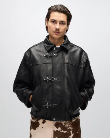 Man wearing a black leather jacket with metal clasp closures and cow-print pants against a light grey studio backdrop.