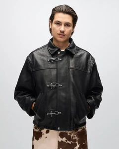 Man wearing a black leather jacket with metal clasp closures and cow-print pants against a light grey studio backdrop.