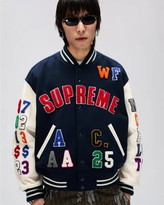 An East Asian man wearing black sunglasses and a navy Supreme varsity jacket with multi-colored patches against a studio backdrop.