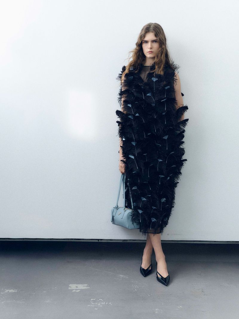 A woman wearing a black feather dress and black heels holds a blue handbag in a studio setting against a white wall.