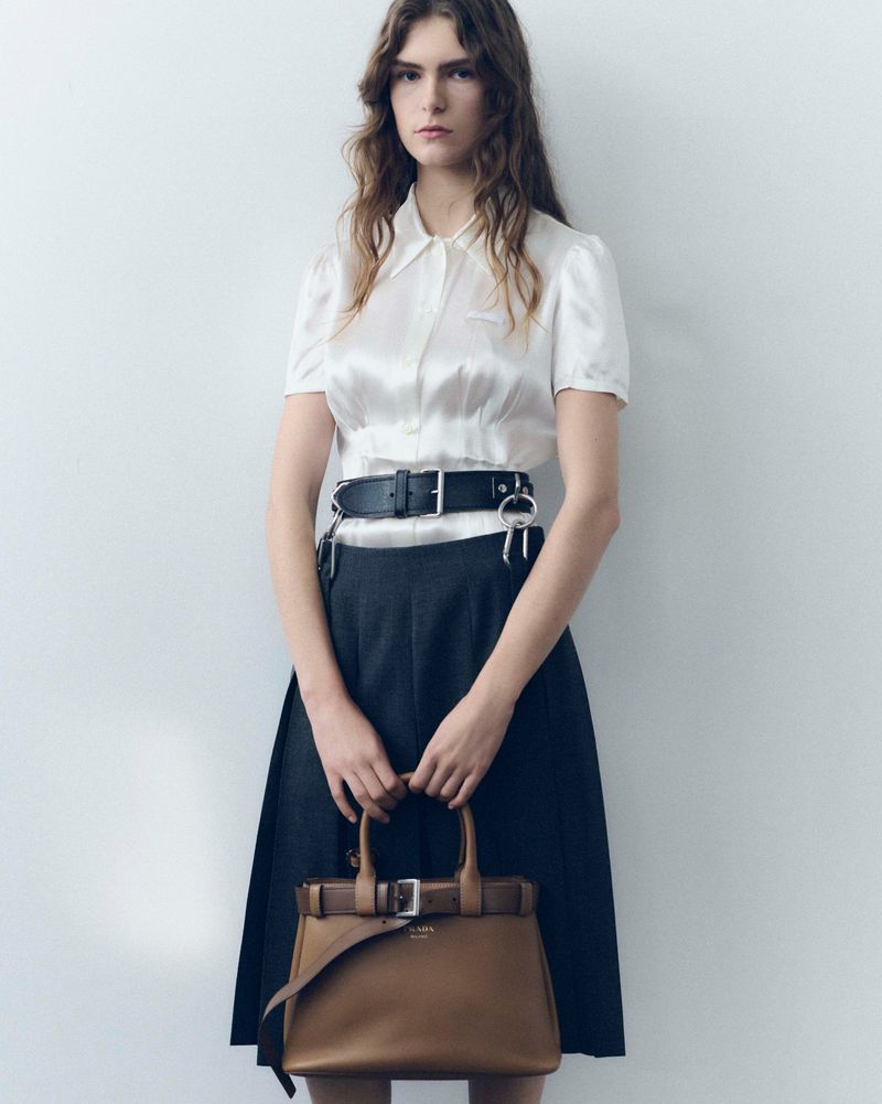 A fashionable woman in a white blouse and dark skirt stands against a white wall, holding a brown leather handbag with buckles.