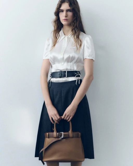 A fashionable woman in a white blouse and dark skirt stands against a white wall, holding a brown leather handbag with buckles.