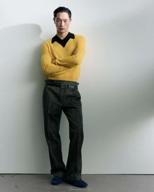 A young adult Asian male stands against a neutral wall, posing in a bright yellow sweater and dark grey trousers with a dark belt and shoes.