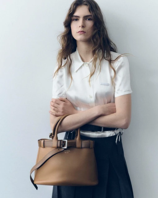 Female fashion model poses indoors with a brown leather Prada handbag, wearing a white button-up shirt and dark skirt against a wall.