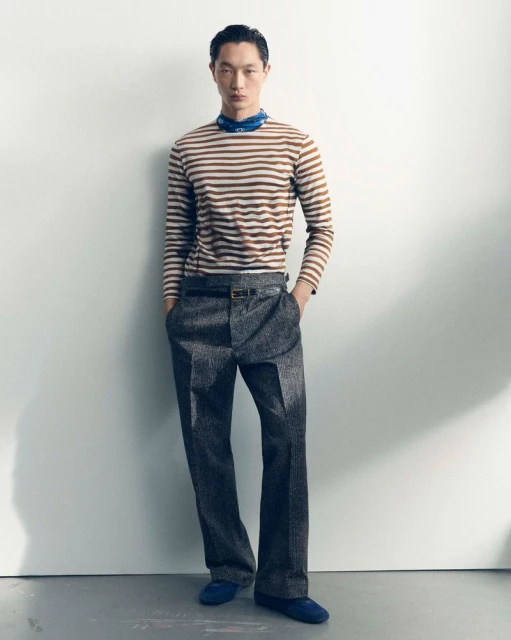 Full-length studio shot: an adult Asian man in a striped long-sleeve shirt and grey trousers poses with hands in pockets, looking at camera.