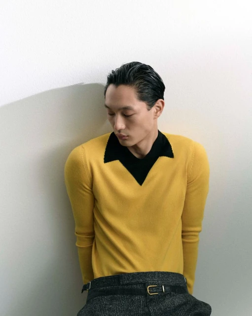 Portrait of a young Asian man in a yellow v-neck sweater, posing indoors against a white wall with his head turned slightly down.