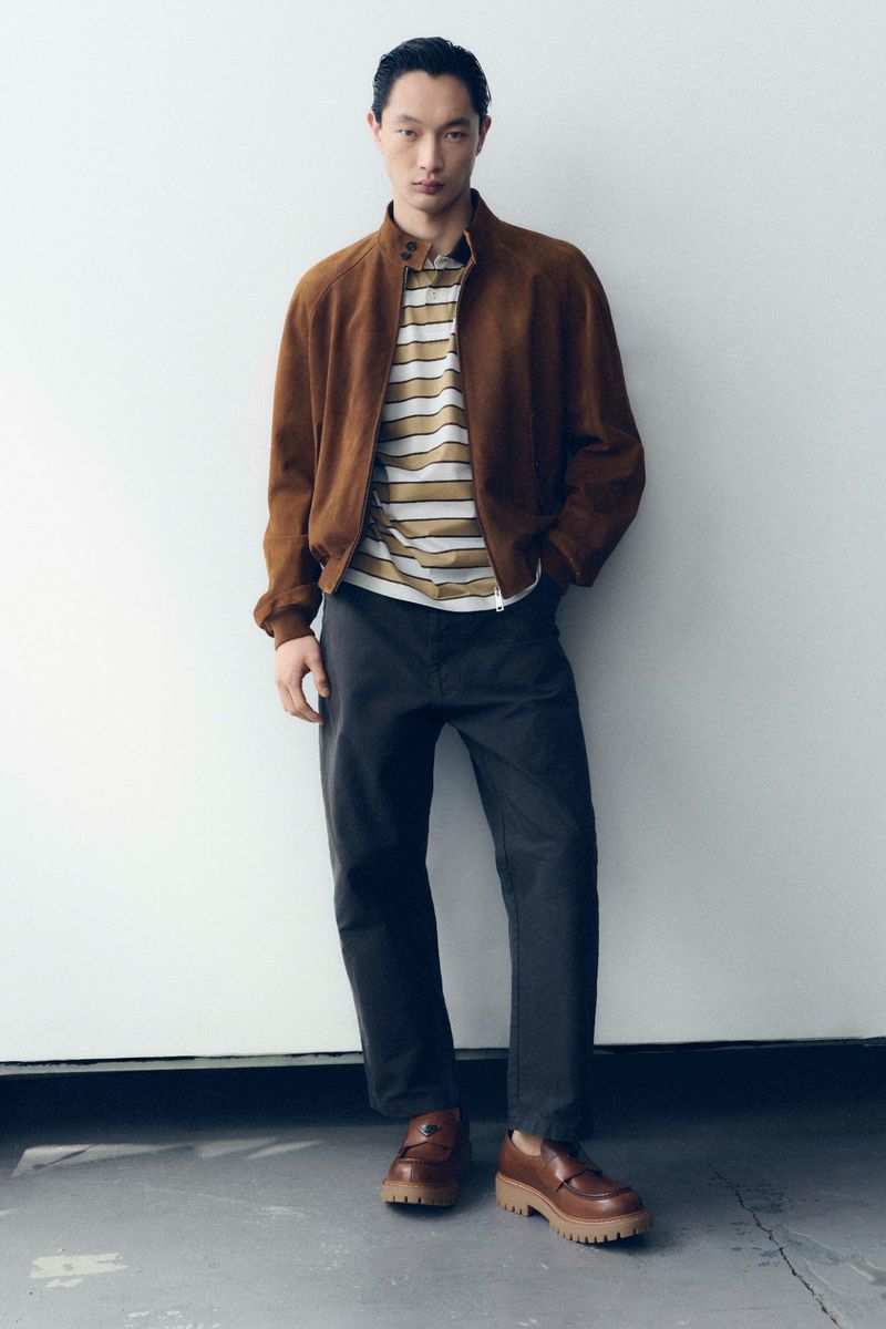 A male model standing against a white wall wearing a brown suede jacket, striped polo shirt, charcoal trousers, and chunky brown loafers.