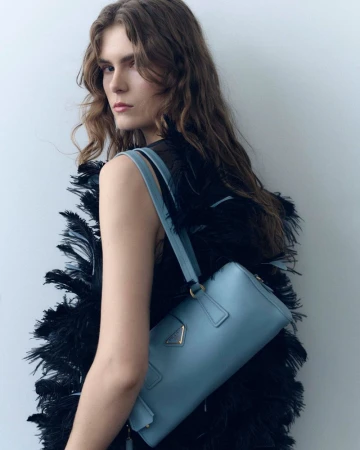 Fashionable woman with long wavy hair wears a black feathered top and poses with a light blue Prada handbag in a studio setting.
