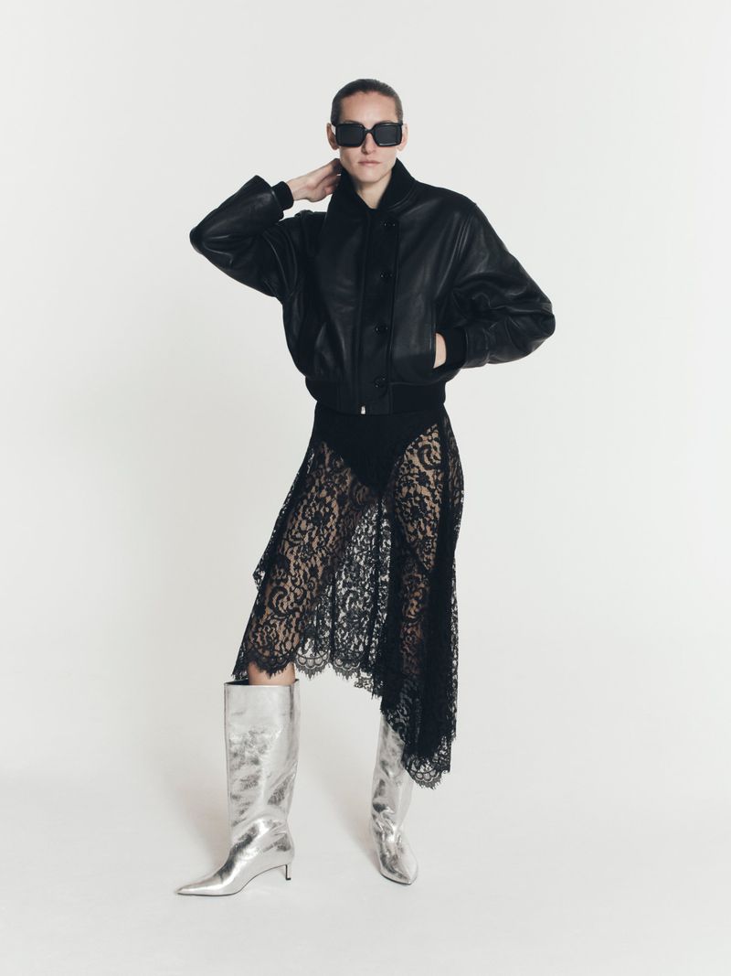 An adult woman in a black leather bomber jacket, lace skirt, and silver boots poses in a full shot against a white backdrop.