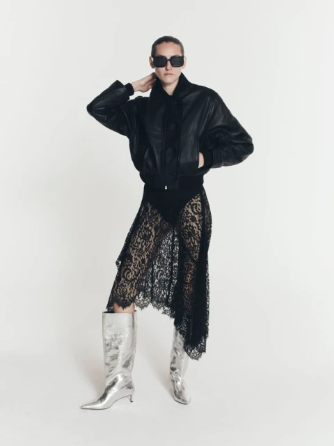 An adult woman in a black leather bomber jacket, lace skirt, and silver boots poses in a full shot against a white backdrop.