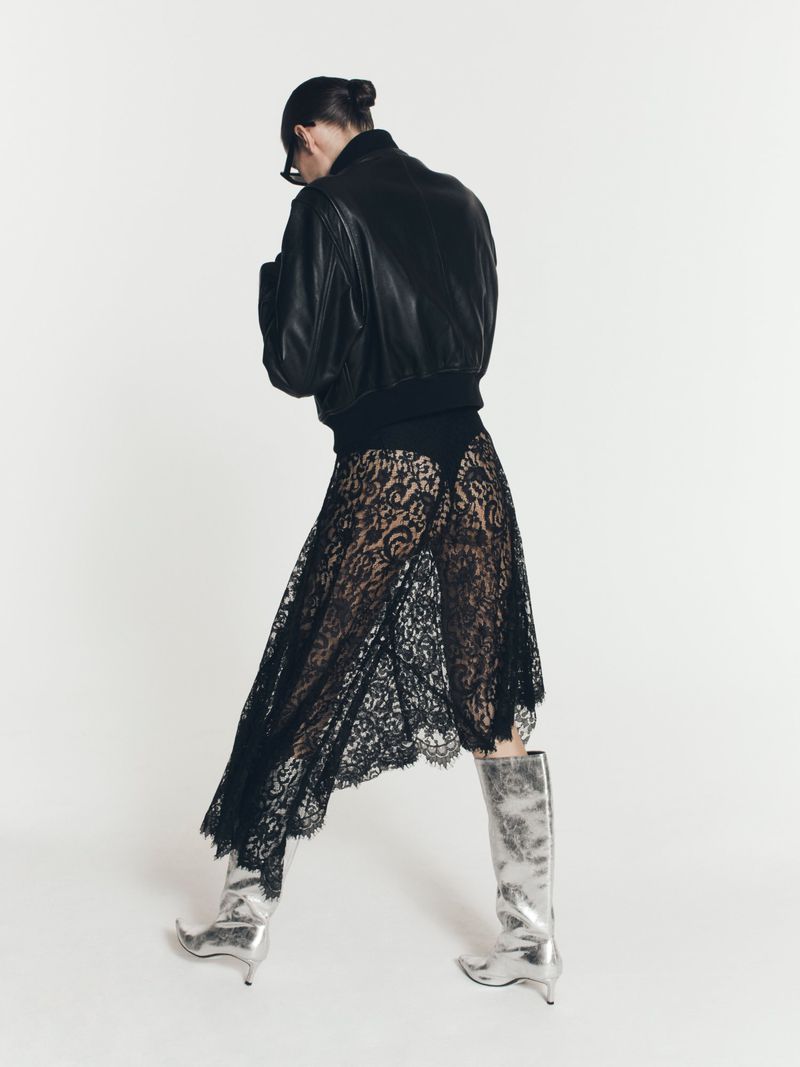 Woman in black bomber jacket, lace skirt, and silver boots poses in a studio with a white backdrop, showcasing a blend of edgy and elegant style.