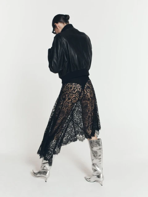 Woman in black bomber jacket, lace skirt, and silver boots poses in a studio with a white backdrop, showcasing a blend of edgy and elegant style.