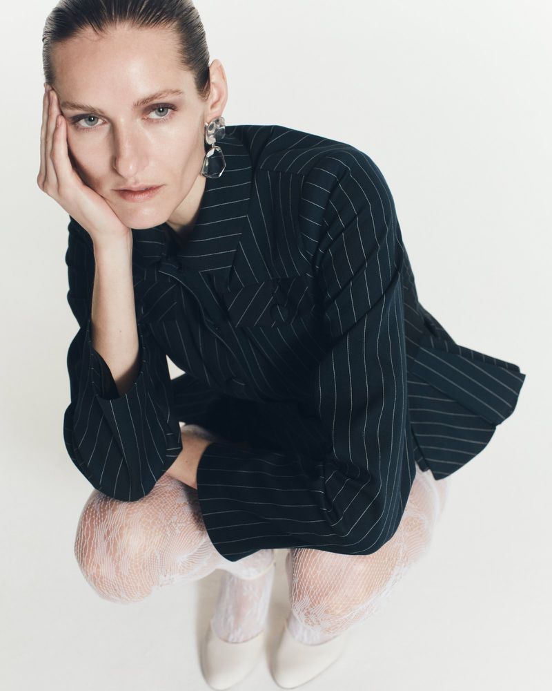 Fashion editorial shot of an adult woman posing in a pinstripe blazer, fishnet tights, and white pumps against a white background in a studio.