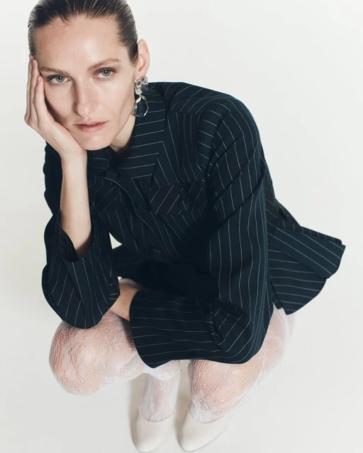 Fashion editorial shot of an adult woman posing in a pinstripe blazer, fishnet tights, and white pumps against a white background in a studio.