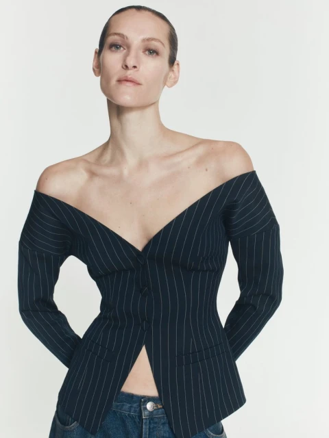 Woman wearing a pinstripe blazer as a top with off-the-shoulder neckline and blue jeans, posing in a studio with a neutral backdrop.