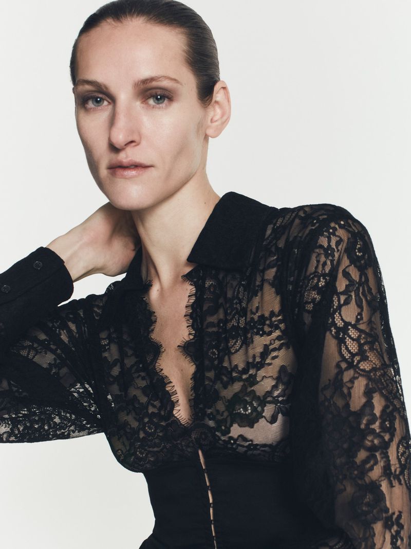 Close-up shot of a woman wearing a black lace top with long sleeves, posing in front of a neutral background in a fashion shot.