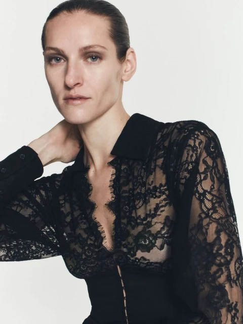 Close-up shot of a woman wearing a black lace top with long sleeves, posing in front of a neutral background in a fashion shot.