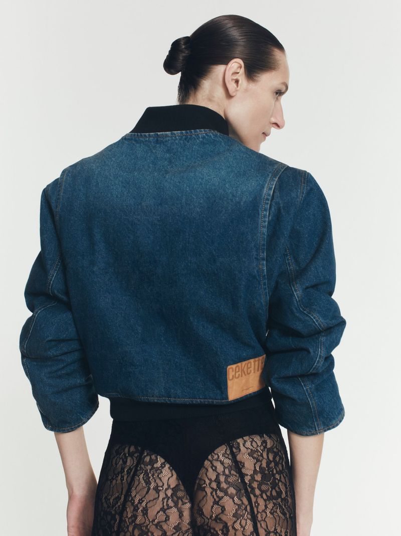 Back view of an adult woman with a topknot wearing a cropped denim jacket, black top, and lace skirt against a white background.