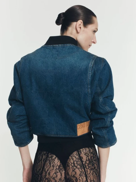 Back view of an adult woman with a topknot wearing a cropped denim jacket, black top, and lace skirt against a white background.