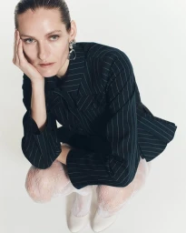 Model Inga Fadina crouching in a dark pinstripe blazer and white lace tights against a clean white seamless studio background.