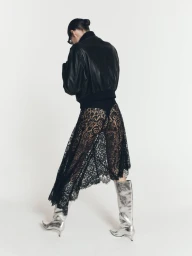 Model from behind wearing a black leather jacket, sheer black lace skirt, and metallic silver knee-high boots on a white backdrop.