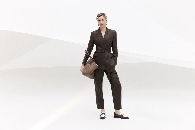 Woman poses in a brown double-breasted suit, floral headscarf, and loafers, holding a tan leather handbag against a gradient white backdrop.