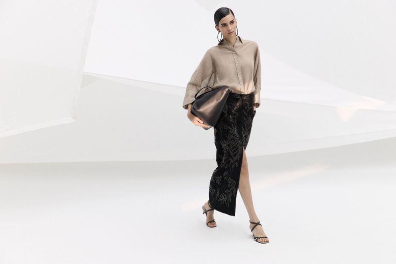 Elegant woman in a beige blouse and black printed skirt walks in a bright white studio holding a black bag, accessorized with hoop earrings.