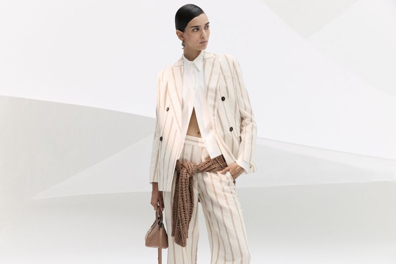 Fashionable woman in a striped linen suit and white shirt holding a handbag in a bright studio setting, showcasing modern elegance.