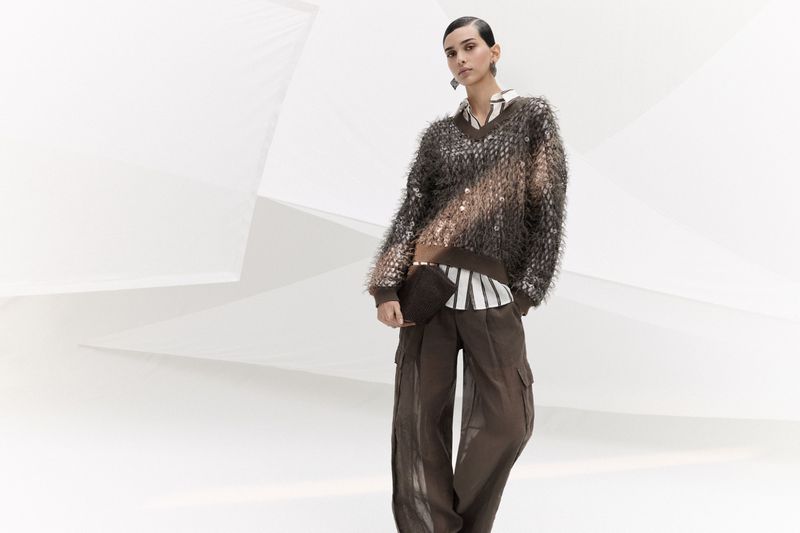 A fashion shot of a woman wearing a brown fuzzy cardigan, a striped shirt, and wide-leg trousers, posing in a minimalist photo studio.