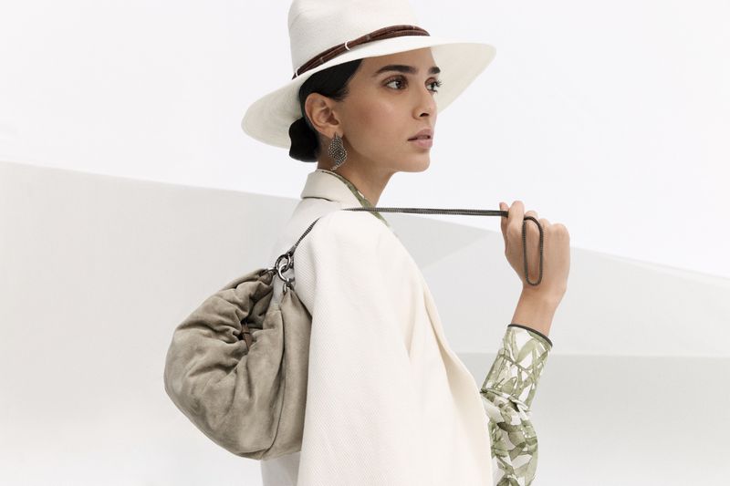 Elegant woman in a cream suit and white hat with a brown band poses indoors with a tan suede slouch bag slung over her shoulder.