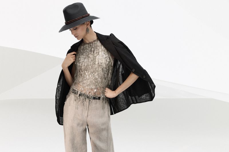 A woman poses in a sparkling top, beige pants, black blazer and hat against a bright white backdrop in a fashion editorial.