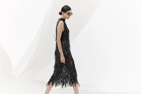Model walking in profile wearing a black sleeveless sequin fringe midi dress and sunglasses against a bright white background.