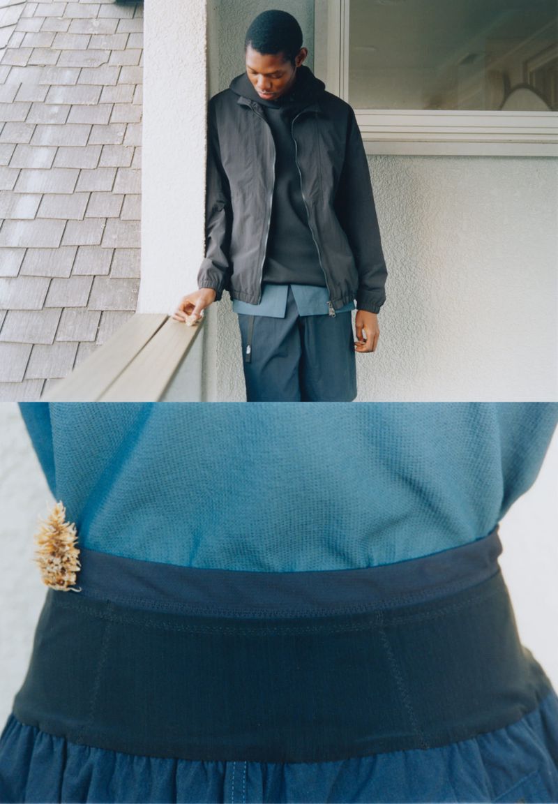 Man in dark jacket and shorts leaning against a wall with tile roof in background, contrasted with torso of woman in blue top and black bottoms.