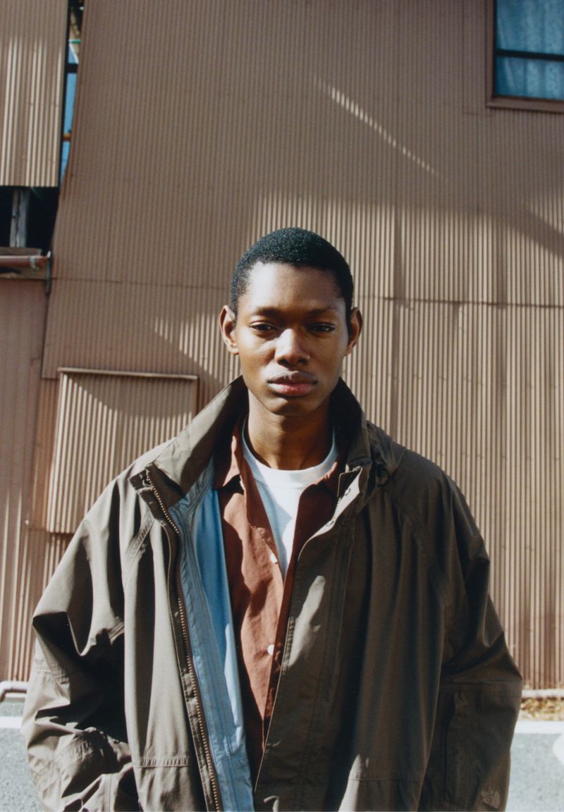 Adult male in earth-tone jacket poses outdoors against corrugated metal wall. Brown shirt peeks beneath blue-trimmed collar. Neutral light.
