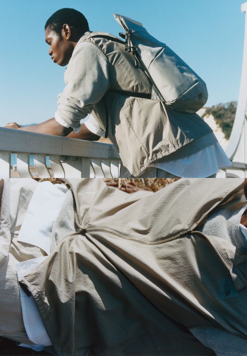 A man with a backpack leans on a railing, dressed in neutral colors, against a light blue sky with an island visible in the distance.