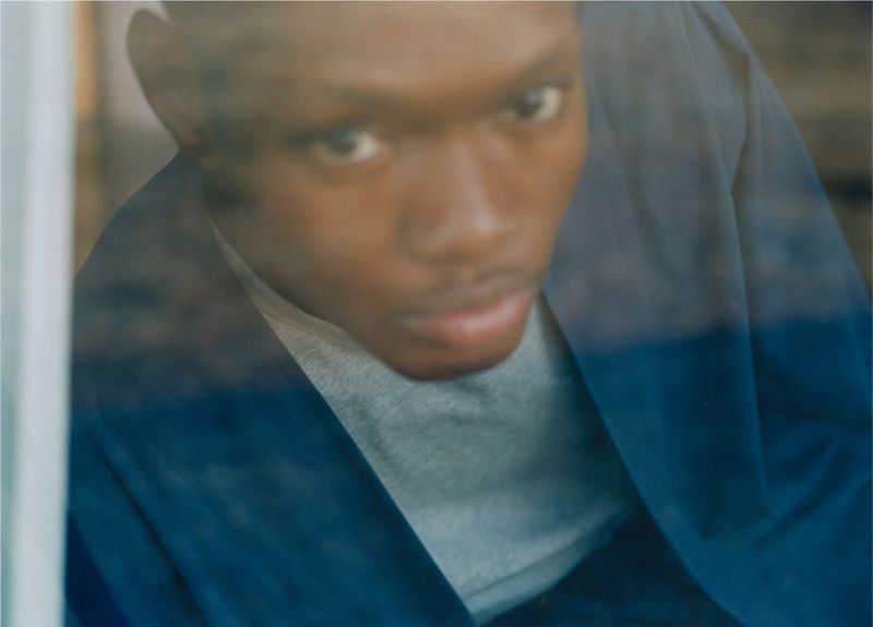Portrait of a young Black man gazing through a glass window, wearing a gray shirt and blue jacket, with soft natural lighting.